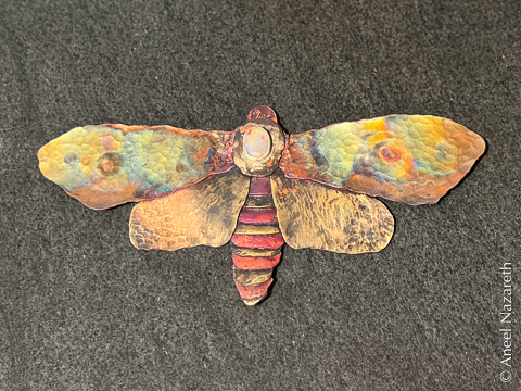 The final version has many colors of oxides on the wings, intended to evoke an ocular pattern. There is a red-orange opal in the center